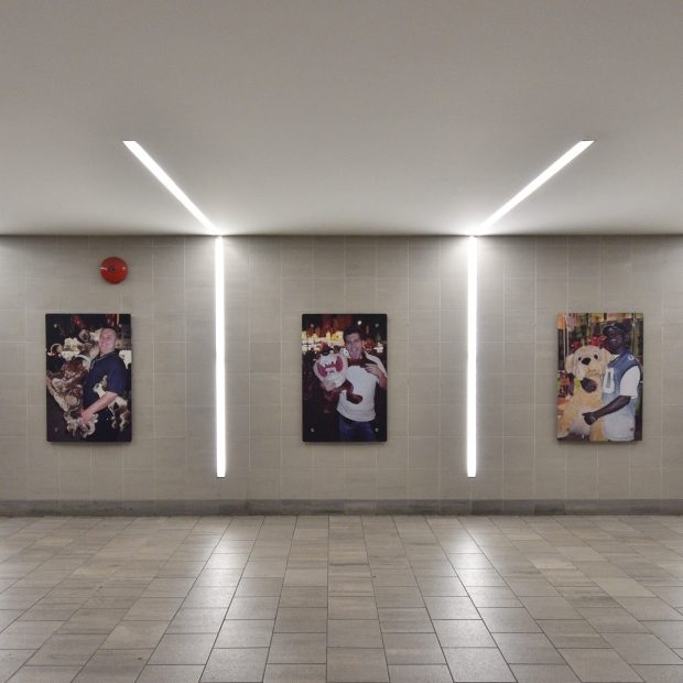 A series of colour, portrait style photographs are installed on a white tile wall inside the SkyTrain Station.