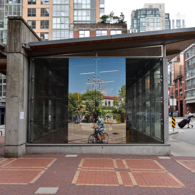 A colour photographic print is presented on the glass face of the SkyTrain station facade. Around the station, the surrounding environment (condo buildings, cars on the street, bricked pathways, grey sky) can be seen.